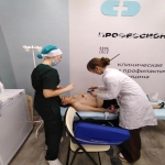 На изображении три человека: один сидит на стуле, другой стоит рядом, третий в белом халате склонился над сидящим. На заднем плане баннер с текстом.
