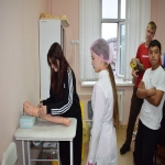 На изображении несколько человек в помещении. Одна девушка ставит укол, другая стоит рядом, остальные наблюдают.