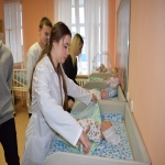 На изображении женщина в белом халате, вероятно, врач или медсестра, работает с пациентом или манекеном. Вокруг неё несколько человек.