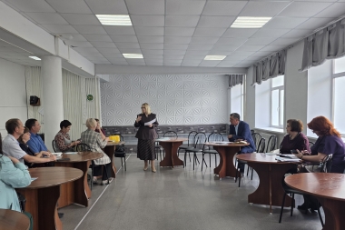 В светлом конференц-зале проходит рабочее совещание: в центре помещения стоит женщина в деловом костюме и с документами в руках, обращаясь к собравшимся. Участники встречи расположились за круглыми деревянными столами по обе стороны зала, внимательно слушая выступающую.