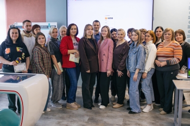 На групповом фото запечатлена большая компания людей, преимущественно женщин, позирующих в современном учебном или выставочном пространстве. На заднем плане виден большой экран с логотипом «ЗДРАВ», а некоторые участники группы держат в руках сертификаты или папки, что указывает на завершение какого-то образовательного или профессионального мероприятия.