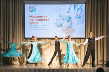На сцене проходит танцевальный номер в рамках торжественного собрания, посвящённого Дню медицинского работника. Артисты в чёрных костюмах и воздушных голубых платьях исполняют парный танец на фоне большого экрана с надписью «Медицина начинается здесь».