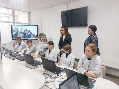 В комнате за столами сидят люди с ноутбуками, рядом стоят преподаватель. На стене висит телевизор.