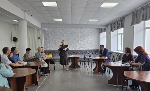 В светлом конференц-зале проходит рабочее совещание: в центре помещения стоит женщина в деловом костюме и с документами в руках, обращаясь к собравшимся. Участники встречи расположились за круглыми деревянными столами по обе стороны зала, внимательно слушая выступающую.