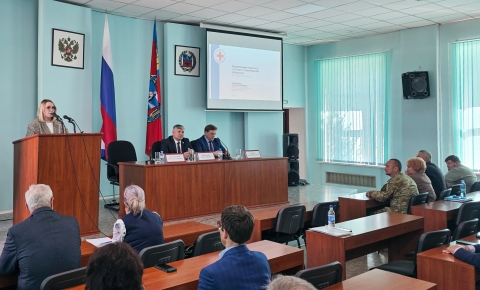 В зале проходит официальное мероприятие: женщина выступает с речью за трибуной, а за длинным столом сидят двое мужчин с микрофонами и табличками с именами. В зале присутствуют слушатели, на стене видны флаги России и региона, а также проекция презентации с заголовком «Реализация проекта „Готов к санитарной обороне“».