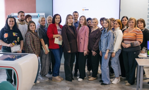 На групповом фото запечатлена большая компания людей, преимущественно женщин, позирующих в современном учебном или выставочном пространстве. На заднем плане виден большой экран с логотипом «ЗДРАВ», а некоторые участники группы держат в руках сертификаты или папки, что указывает на завершение какого-то образовательного или профессионального мероприятия.