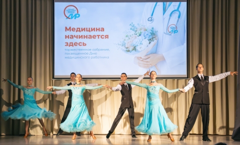 На сцене проходит танцевальный номер в рамках торжественного собрания, посвящённого Дню медицинского работника. Артисты в чёрных костюмах и воздушных голубых платьях исполняют парный танец на фоне большого экрана с надписью «Медицина начинается здесь».