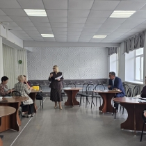 В светлом конференц-зале проходит рабочее совещание: в центре помещения стоит женщина в деловом костюме и с документами в руках, обращаясь к собравшимся. Участники встречи расположились за круглыми деревянными столами по обе стороны зала, внимательно слушая выступающую.
