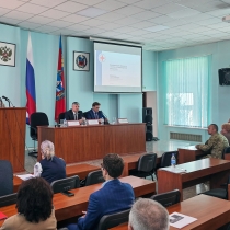 В зале проходит официальное мероприятие: женщина выступает с речью за трибуной, а за длинным столом сидят двое мужчин с микрофонами и табличками с именами. В зале присутствуют слушатели, на стене видны флаги России и региона, а также проекция презентации с заголовком «Реализация проекта „Готов к санитарной обороне“».