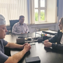 В светлом кабинете за большим столом сидят двое собеседников: мужчина в серой футболке и медицинской шапочке и женщина в деловом пиджаке в полоску, они внимательно смотрят друг на друга, сложив руки. На заднем плане в кресле расположился ещё один мужчина в светлой рубашке, наблюдающий за диалогом, а за окнами видна зелёная листва.