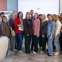 На групповом фото запечатлена большая компания людей, преимущественно женщин, позирующих в современном учебном или выставочном пространстве. На заднем плане виден большой экран с логотипом «ЗДРАВ», а некоторые участники группы держат в руках сертификаты или папки, что указывает на завершение какого-то образовательного или профессионального мероприятия.