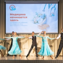 На сцене проходит танцевальный номер в рамках торжественного собрания, посвящённого Дню медицинского работника. Артисты в чёрных костюмах и воздушных голубых платьях исполняют парный танец на фоне большого экрана с надписью «Медицина начинается здесь».