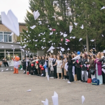 На изображении видна большая группа людей, стоящих на площади. Вокруг растут деревья, на заднем плане виднеется здание.
