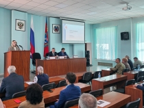 В зале проходит официальное мероприятие: женщина выступает с речью за трибуной, а за длинным столом сидят двое мужчин с микрофонами и табличками с именами. В зале присутствуют слушатели, на стене видны флаги России и региона, а также проекция презентации с заголовком «Реализация проекта „Готов к санитарной обороне“».