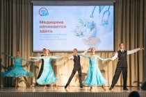 На сцене проходит танцевальный номер в рамках торжественного собрания, посвящённого Дню медицинского работника. Артисты в чёрных костюмах и воздушных голубых платьях исполняют парный танец на фоне большого экрана с надписью «Медицина начинается здесь».
