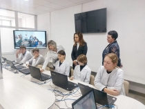 В комнате за столами сидят люди с ноутбуками, рядом стоят преподаватель. На стене висит телевизор.
